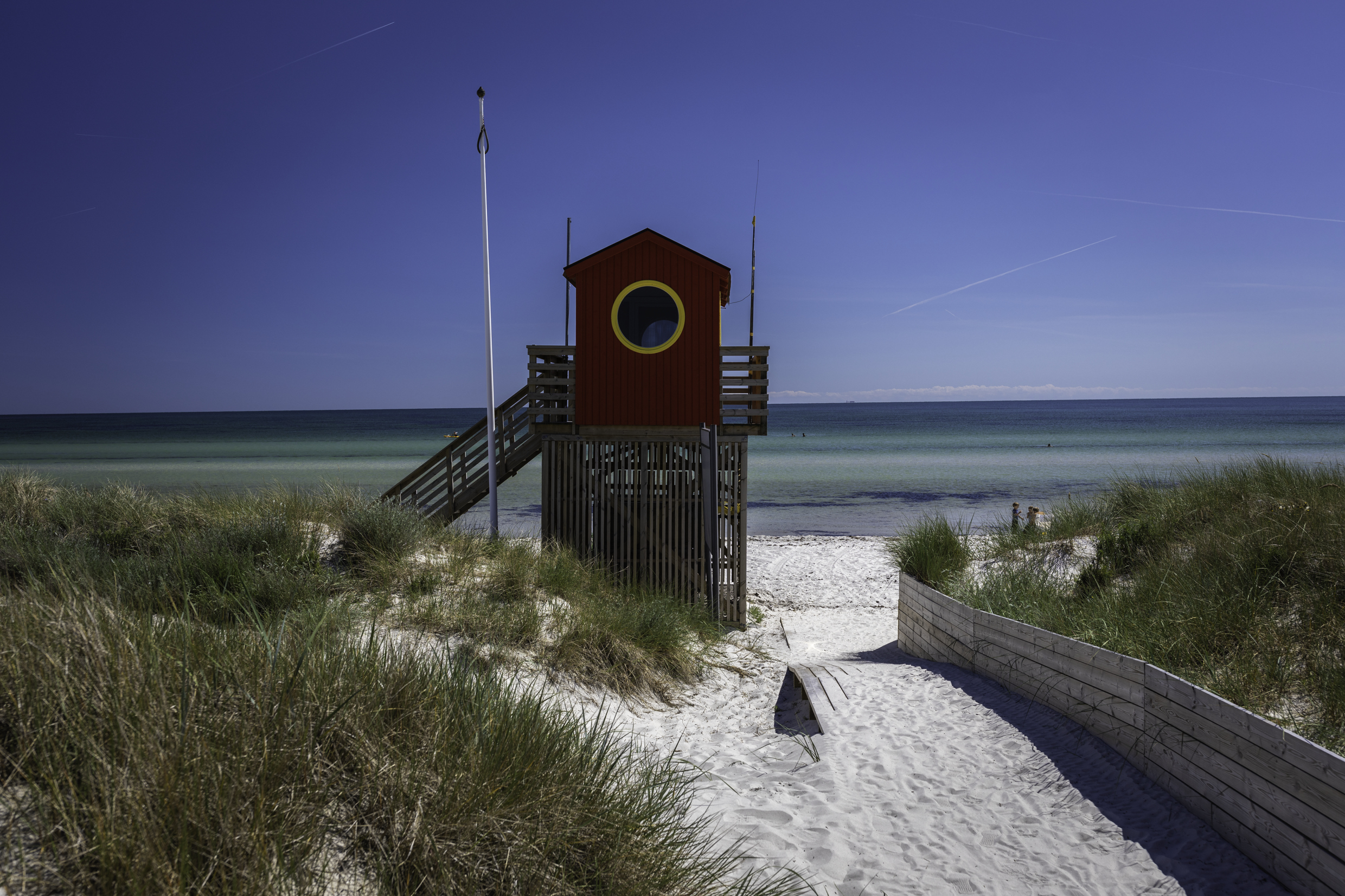 Wooden lifeguard tower with circular window on sandy beach path through dunes, overlooking calm blue sea under twilight sky
