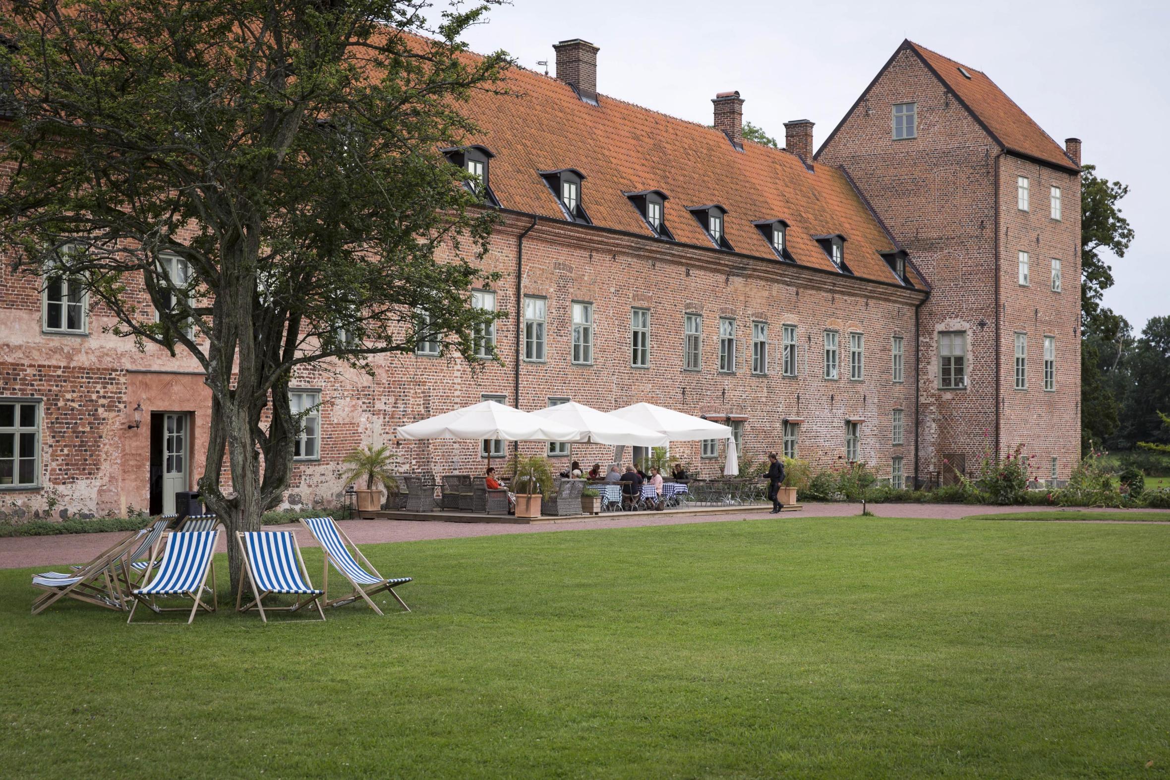 Red-bricked castle with a tower to the right and a tree with blue-striped chairs underneath