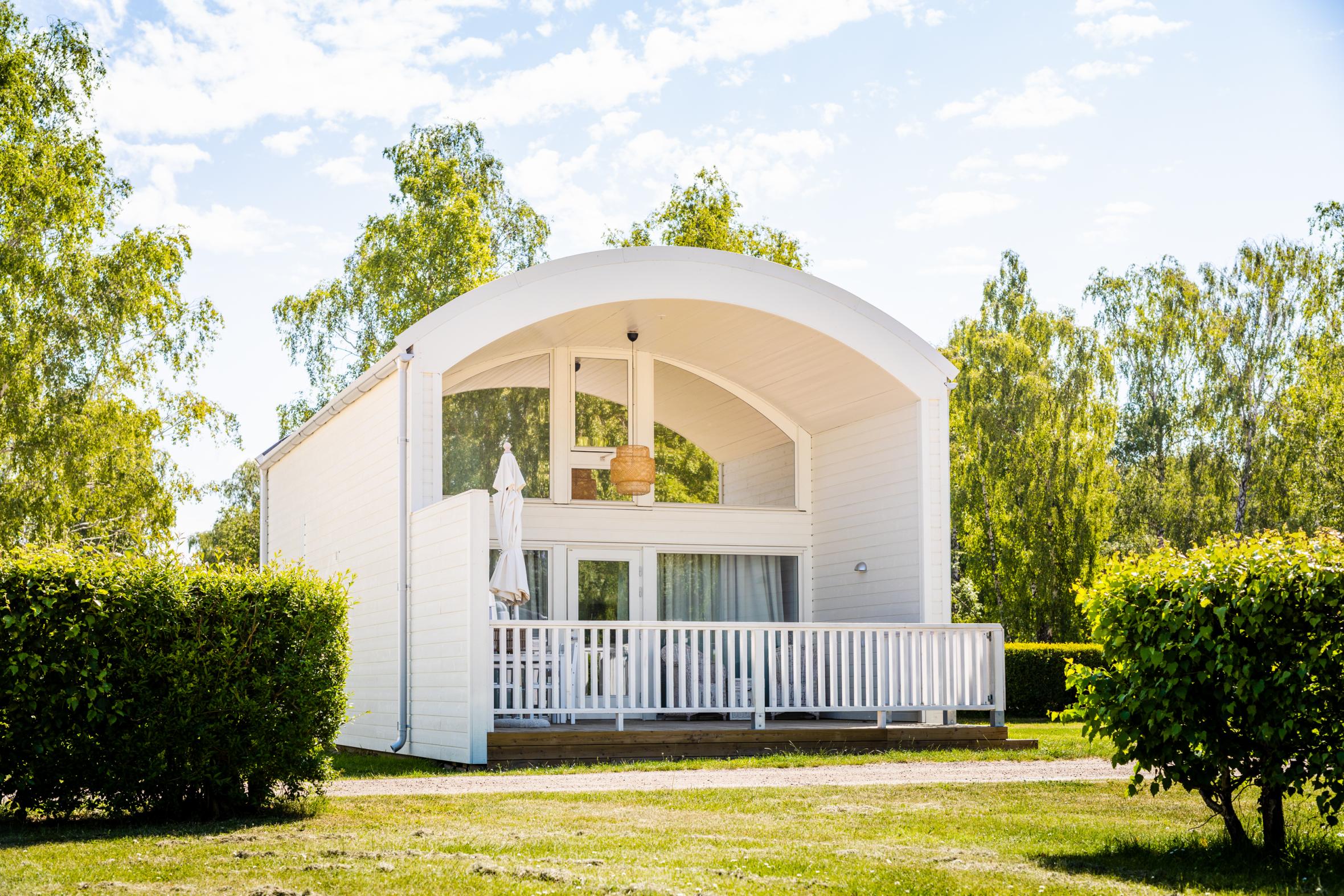 Ein außergewöhnlich gebautes weißes Sommerhäuschen mit gewölbtem Dach und Rasen und Sträuchern vor der Terrasse.