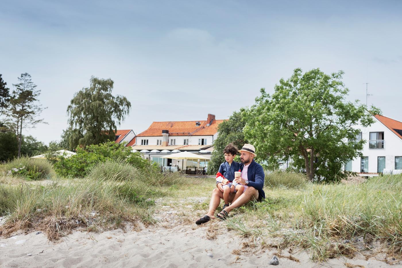 Ein Vater sitzt mit seinem Sohn vor einem großen weißen Haus an einem Sandstrand