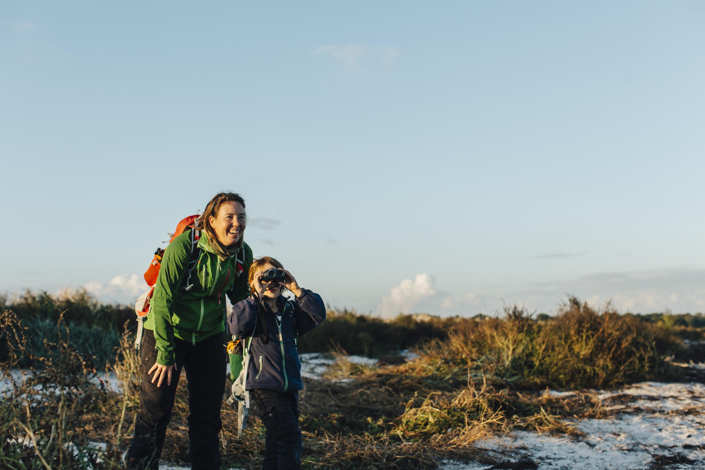 Smiling woman with backpack and child using binoculars in natural landscape, both dressed for outdoor adventure in golden light
