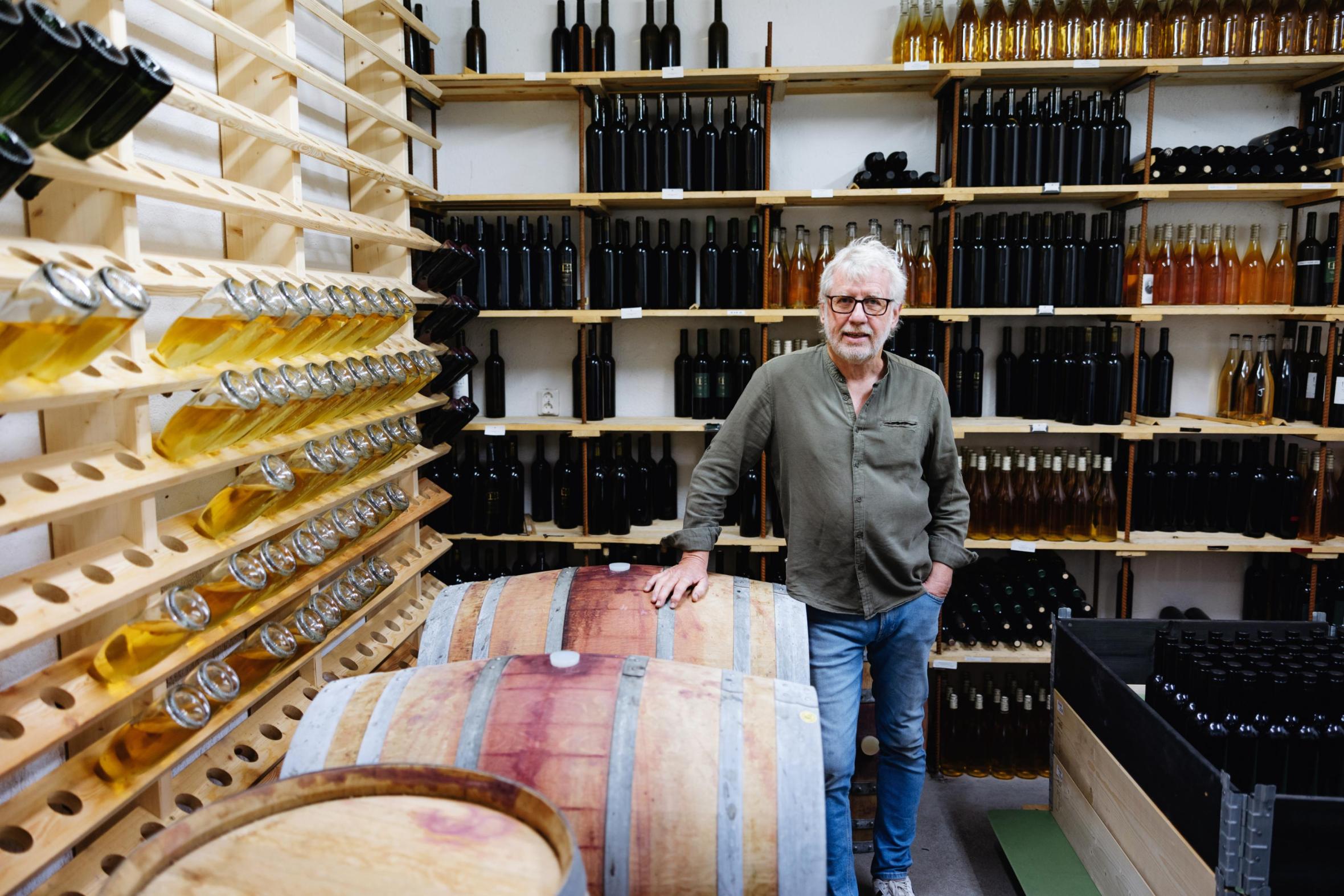 Winemaker with glasses standing beside wooden wine barrel in cellar, surrounded by shelves filled with wine bottles and wooden wine racks