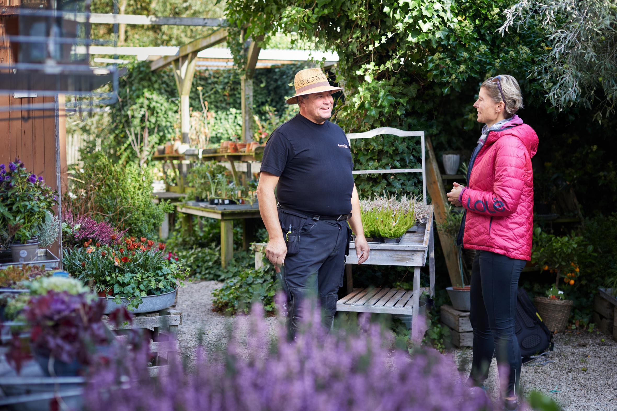 Man in straw hat and woman in jacket having friendly conversation in nursery garden surrounded by potted plants, flowers and garden displays