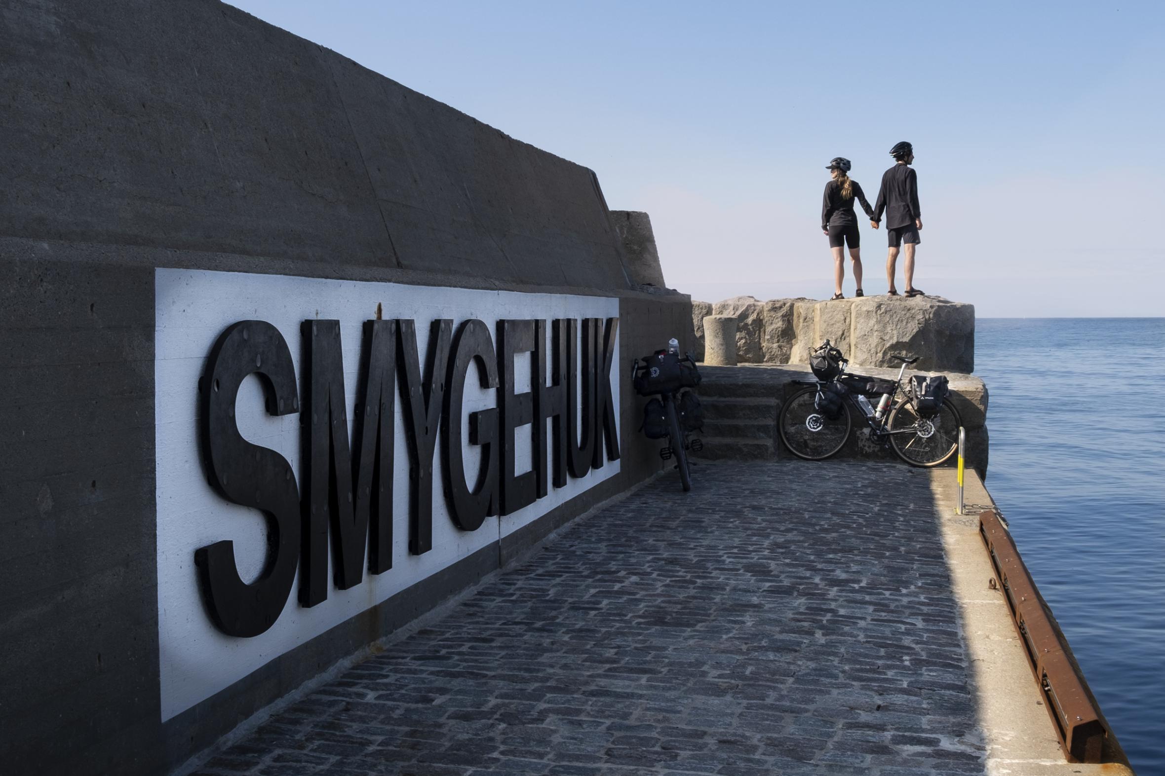 English: Couple holding hands on concrete breakwater with large "SMYGEHUK" sign, bicycles parked nearby, overlooking calm sea at Sweden's southernmost point
