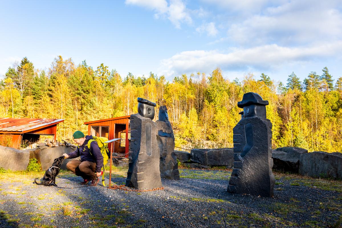Person with dog resting beside large carved stone sculptures in quarry setting, autumn forest backdrop with golden birch trees and clear sky