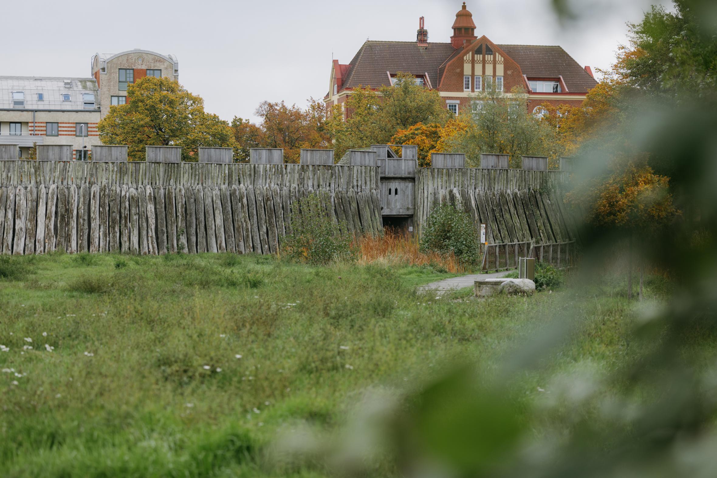 Historic wooden palisade fortification "trelleborg" with watchtower and gate, modern residential buildings visible beyond defensive walls, green parkland in foreground