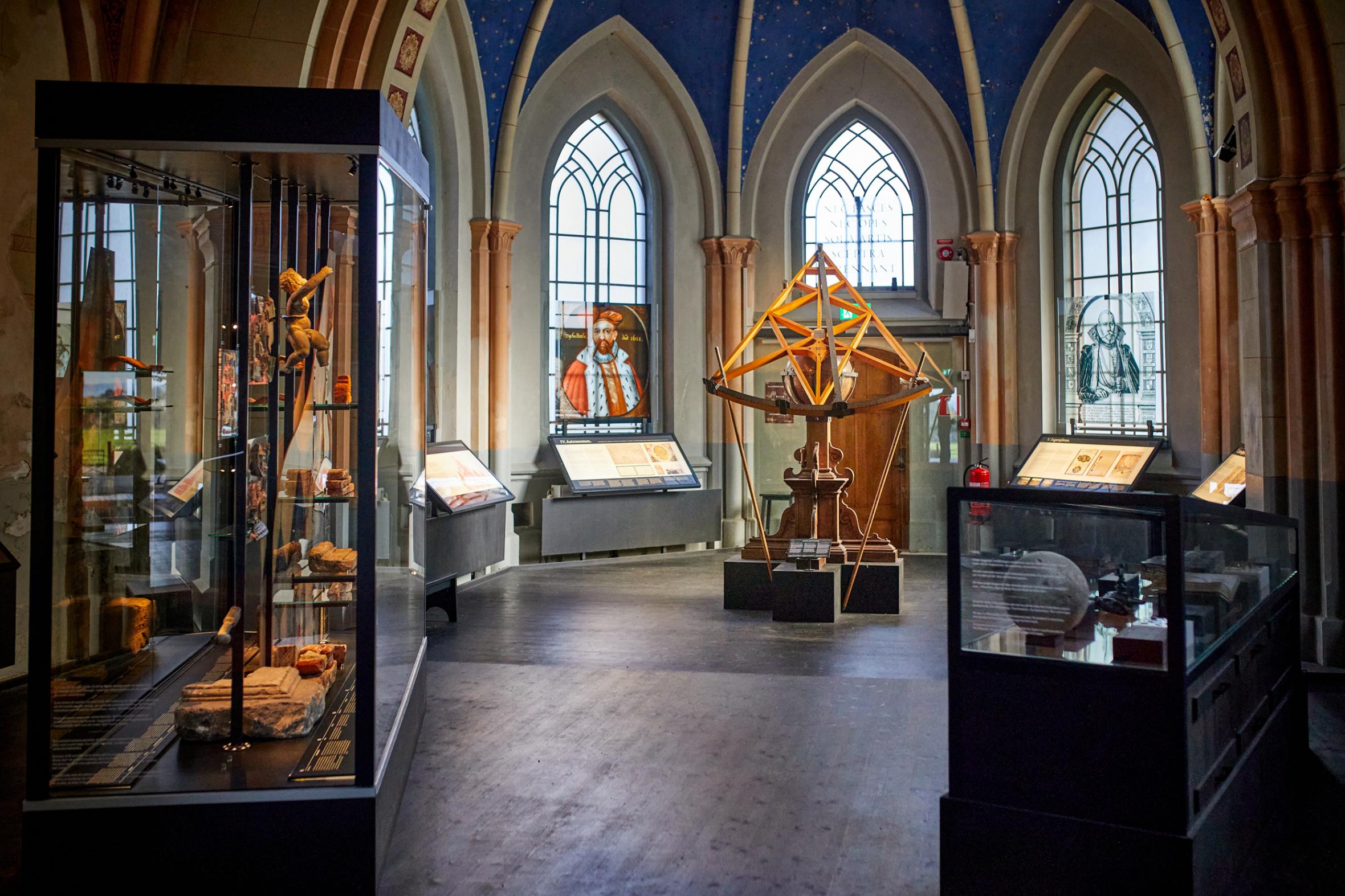English: Museum interior in Gothic cathedral with arched windows, display cases containing historical artifacts, geometric wooden sculpture and stained glass portraits