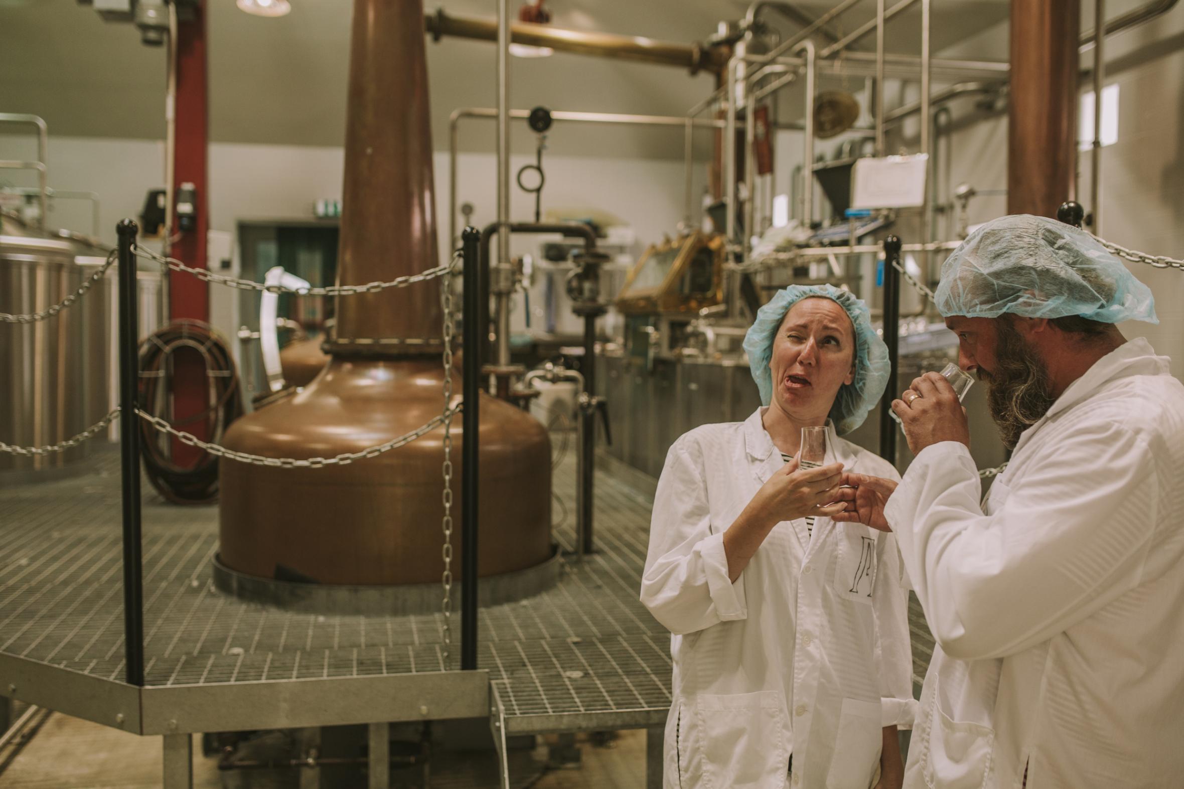 Two people in white lab coats and hair nets tasting samples in modern brewery with copper brewing tanks and industrial equipment