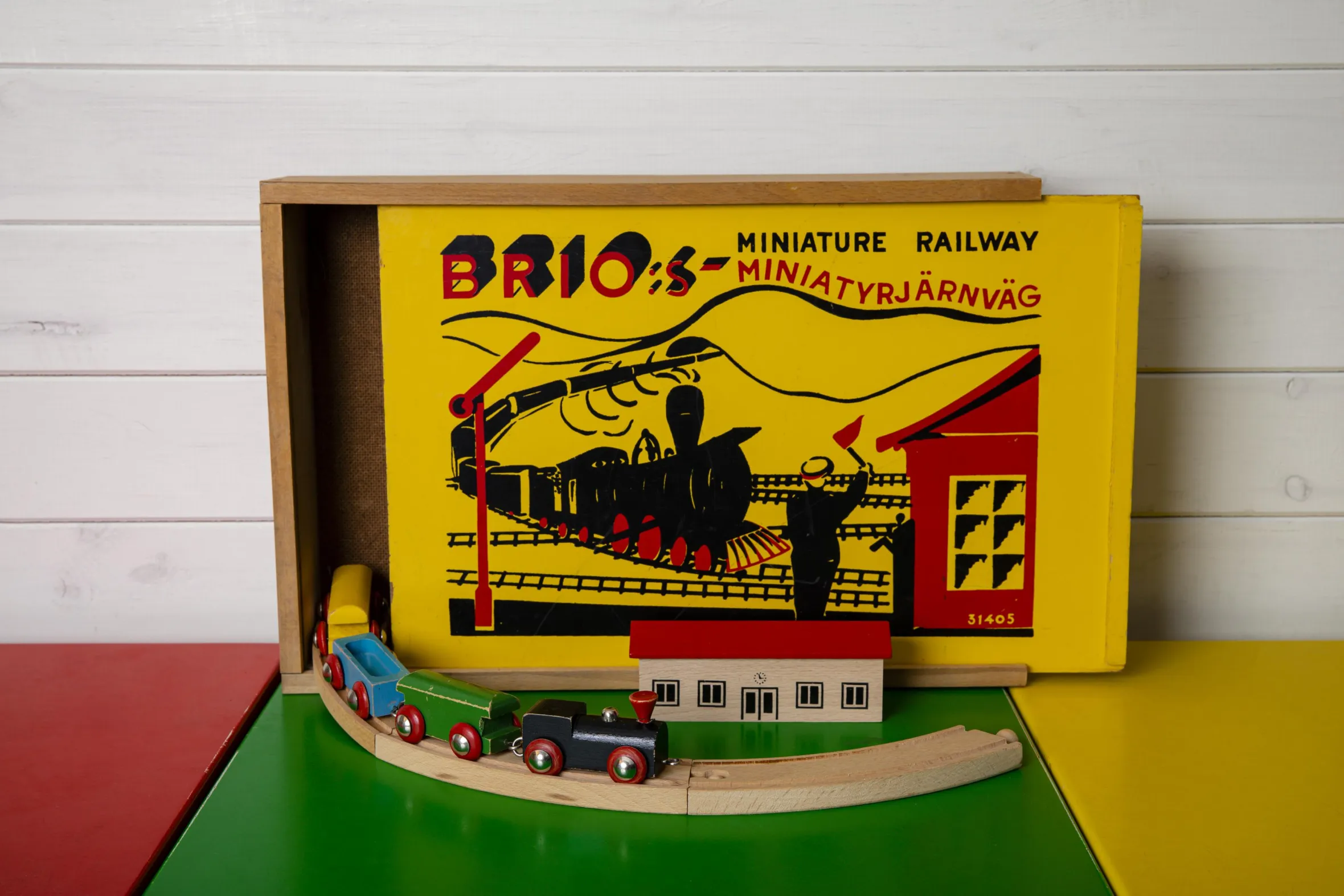 Vintage Brio miniature railway toy set with original box, wooden train and tracks displayed on colorful play mat against white wooden wall