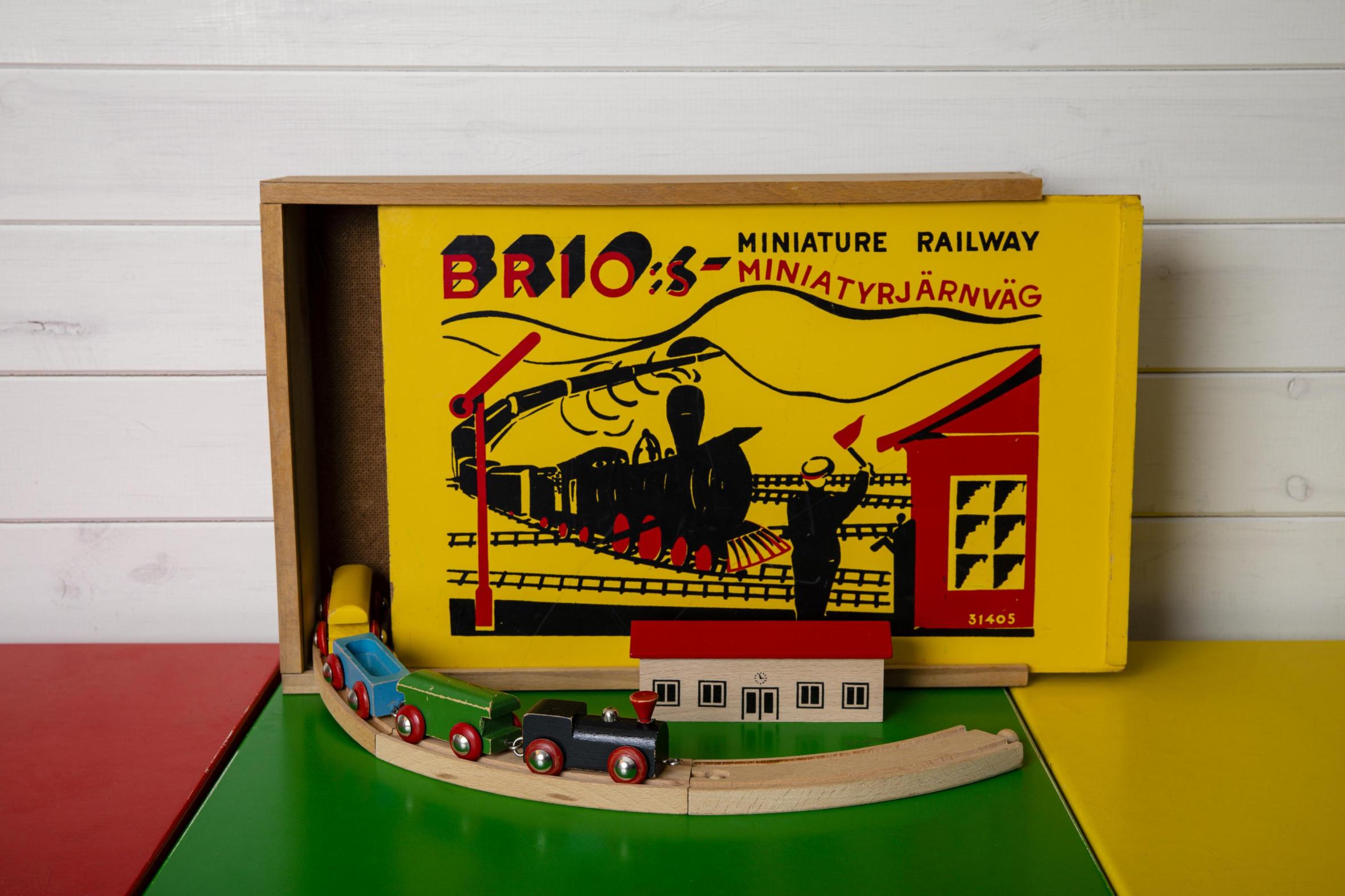 Vintage Brio miniature railway toy set with original box, wooden train and tracks displayed on colorful play mat against white wooden wall