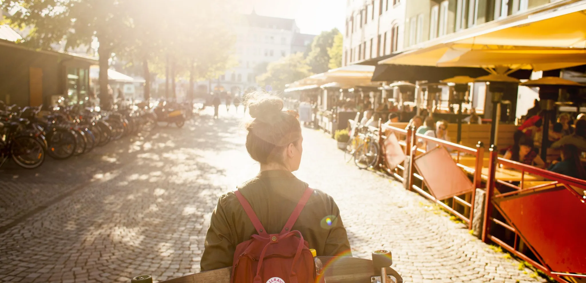 An einem sonnigen Abend geht eine Frau mit Rucksack und Skateboard auf dem Rücken über den Platz Möllevångstorget in Malmö.