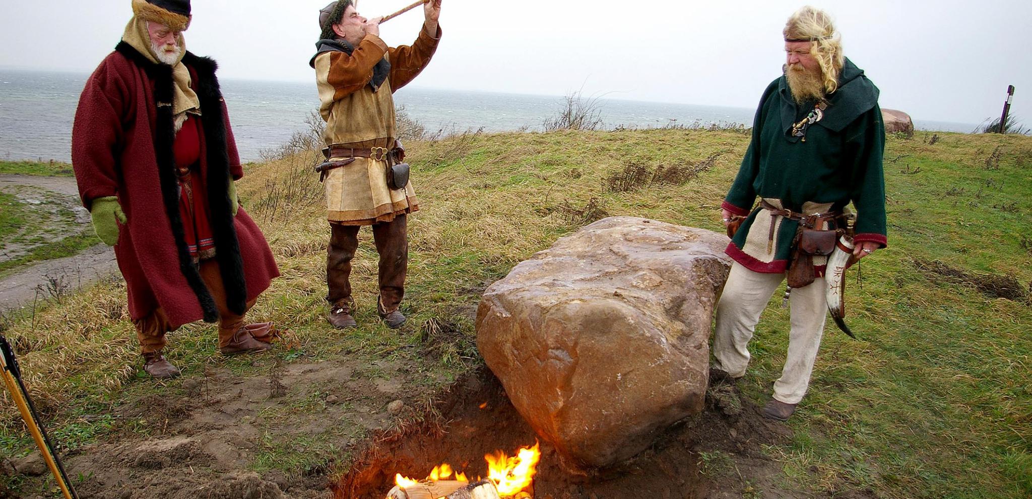 Drei Wikinger stehen um ein Lagerfeuer, einer von ihnen bläst in ein längliches Horn.