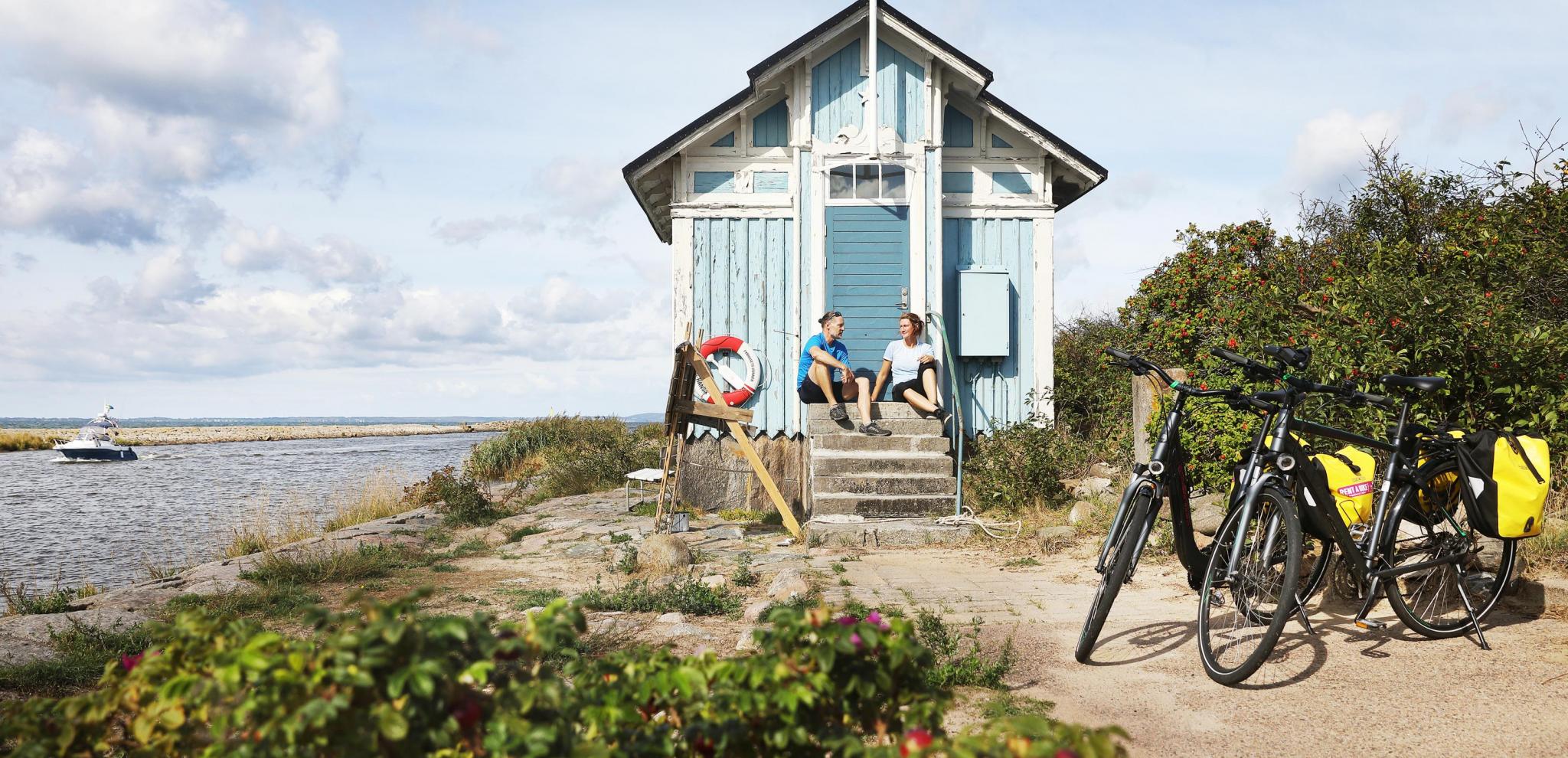 Ein Paar mit Fahrrädern macht eine Pause vor einem hellblauen Strandhäuschen.