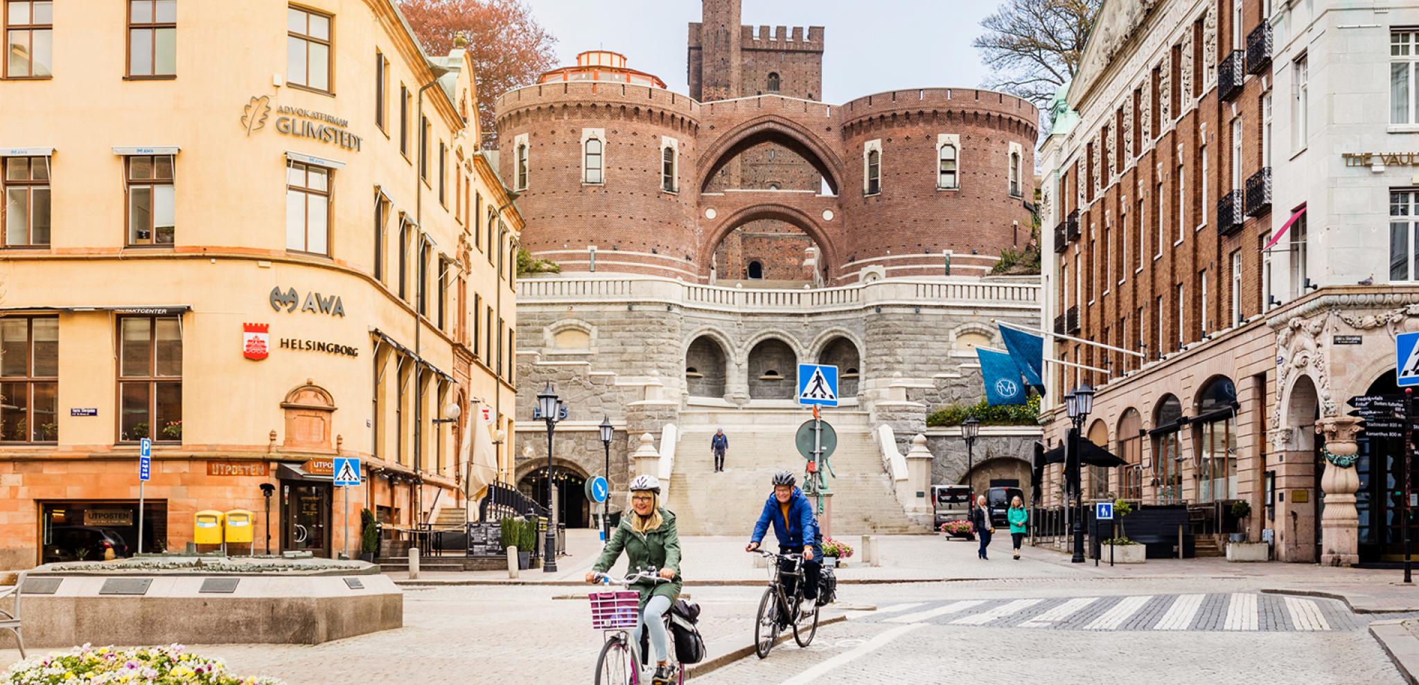 Ein Paar radelt durch das Zentrum von Helsingborg und hat die Treppen zum Turm Kärnan im Hintergrund.