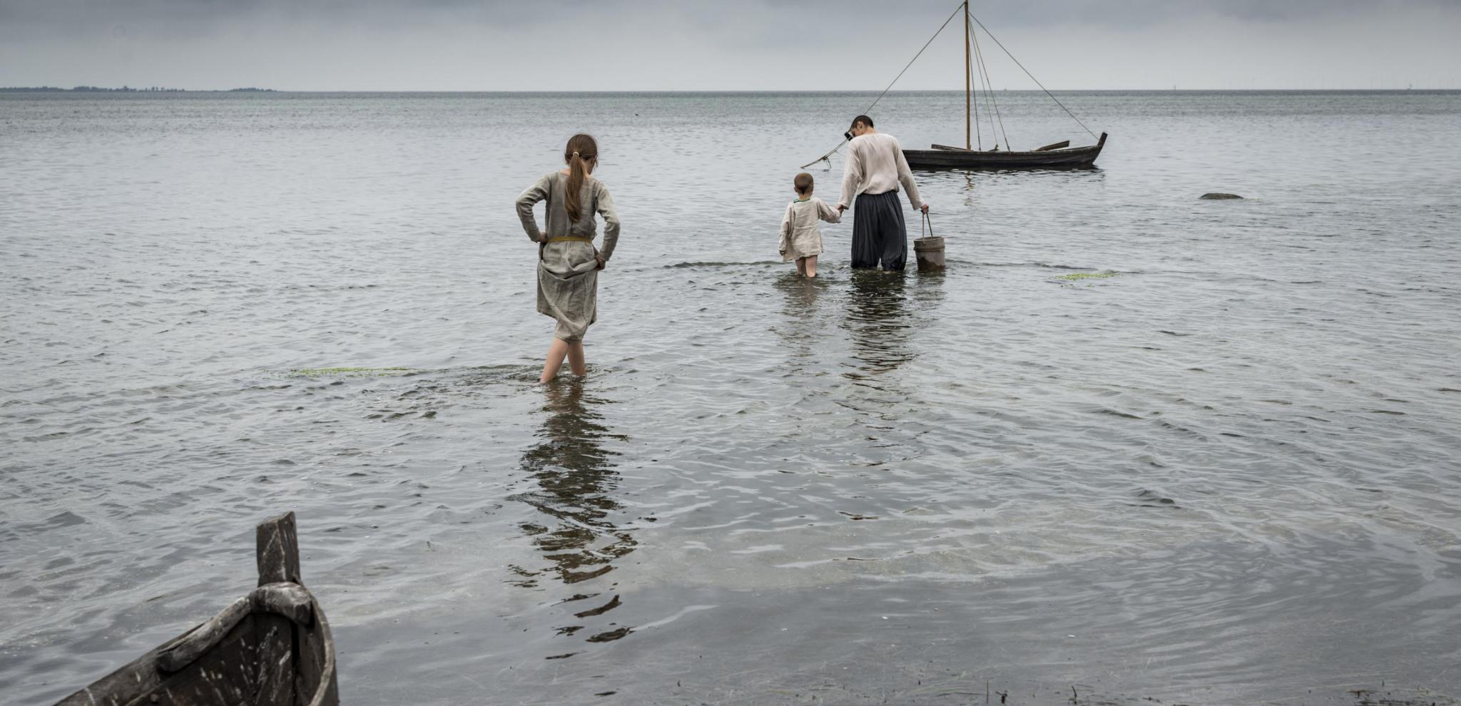 Eine als Wikinger verkleidete Familie watet durch flaches Wasser auf ein Holzboot zu.