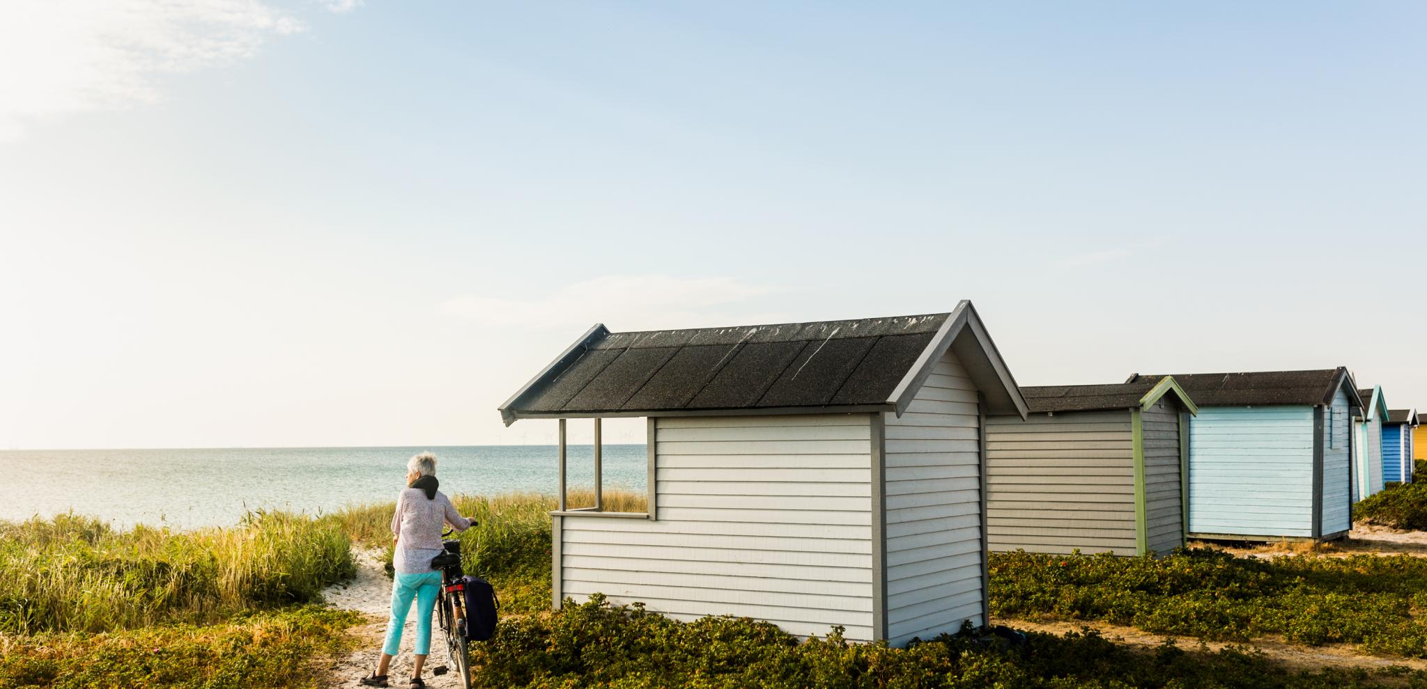 Eine Frau mit einem Fahrrad steht neben einer weißen Strandhütte und blickt auf das Meer.