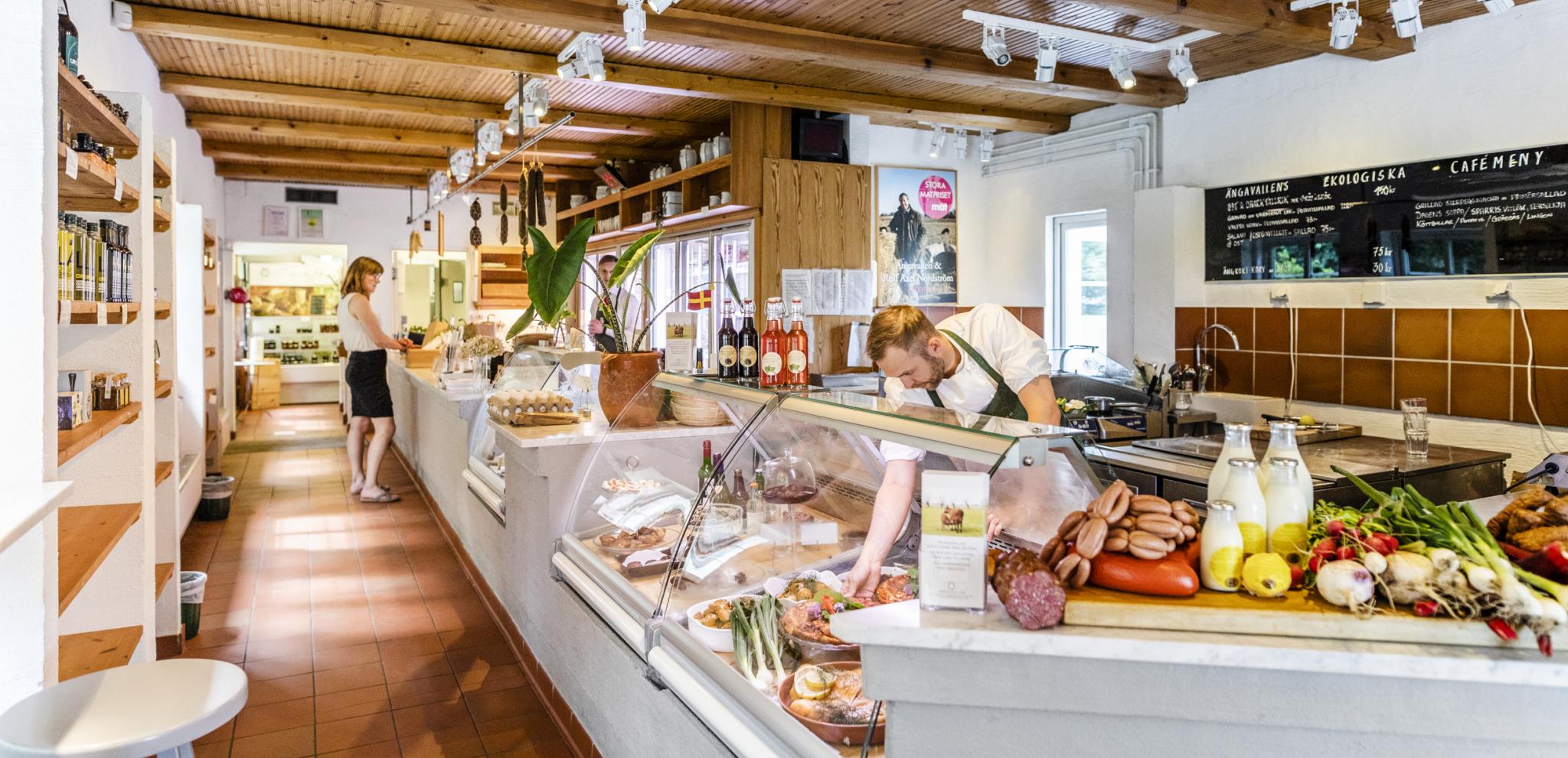 Modern delicatessen interior with exposed wooden ceiling beams, display counter filled with fresh foods, staff serving customers in bright retail space