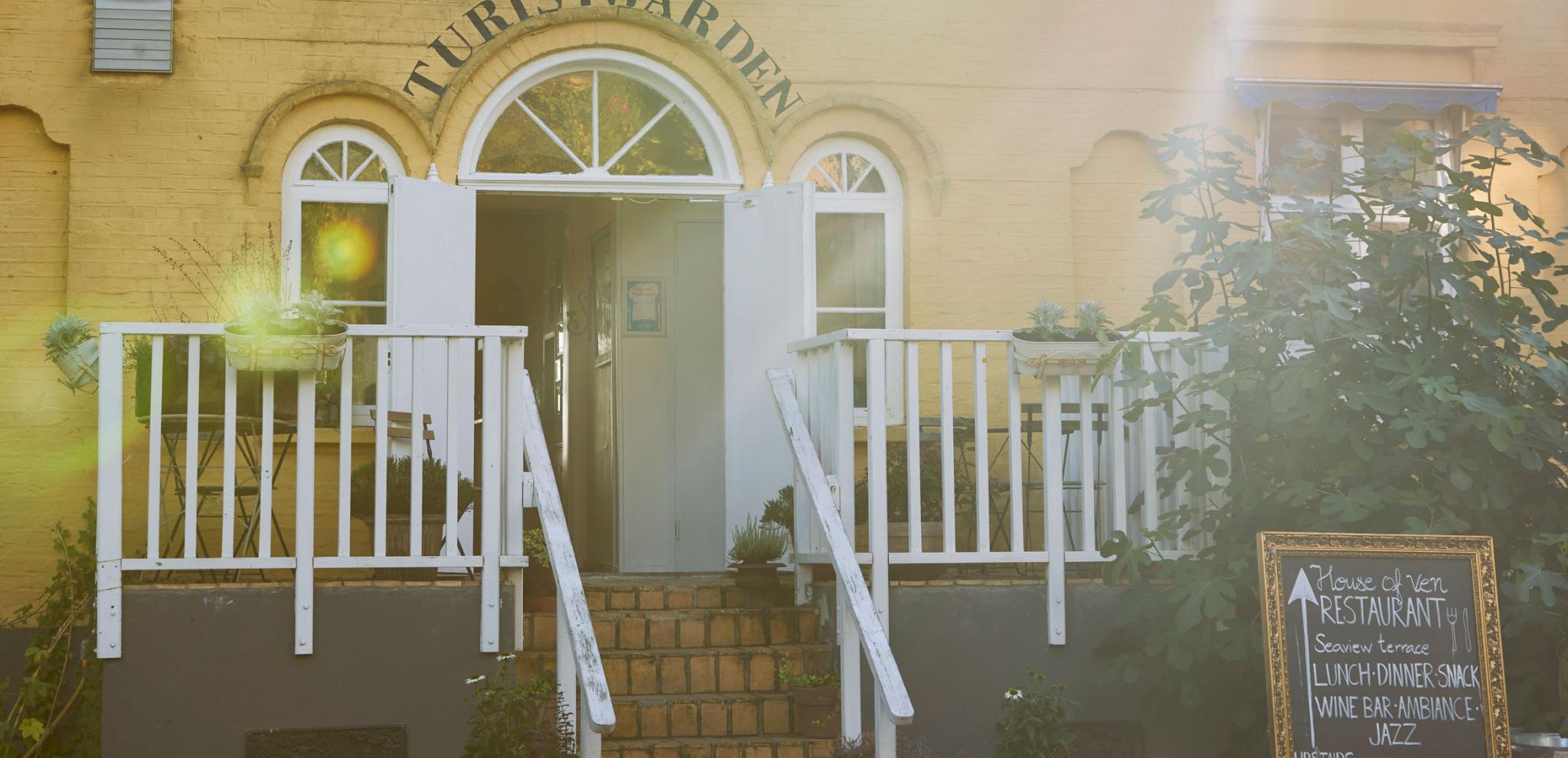 Historic building entrance with "TURISTGÅRDEN" text above arched windows, white wooden stairs and balcony, restaurant menu board visible