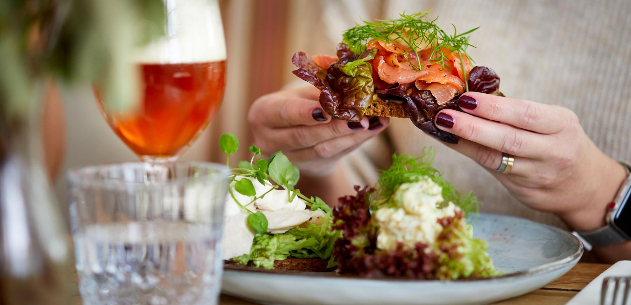 ands holding open-faced sandwich with smoked salmon and herbs, wine glass and plated appetizers on wooden table in restaurant setting
