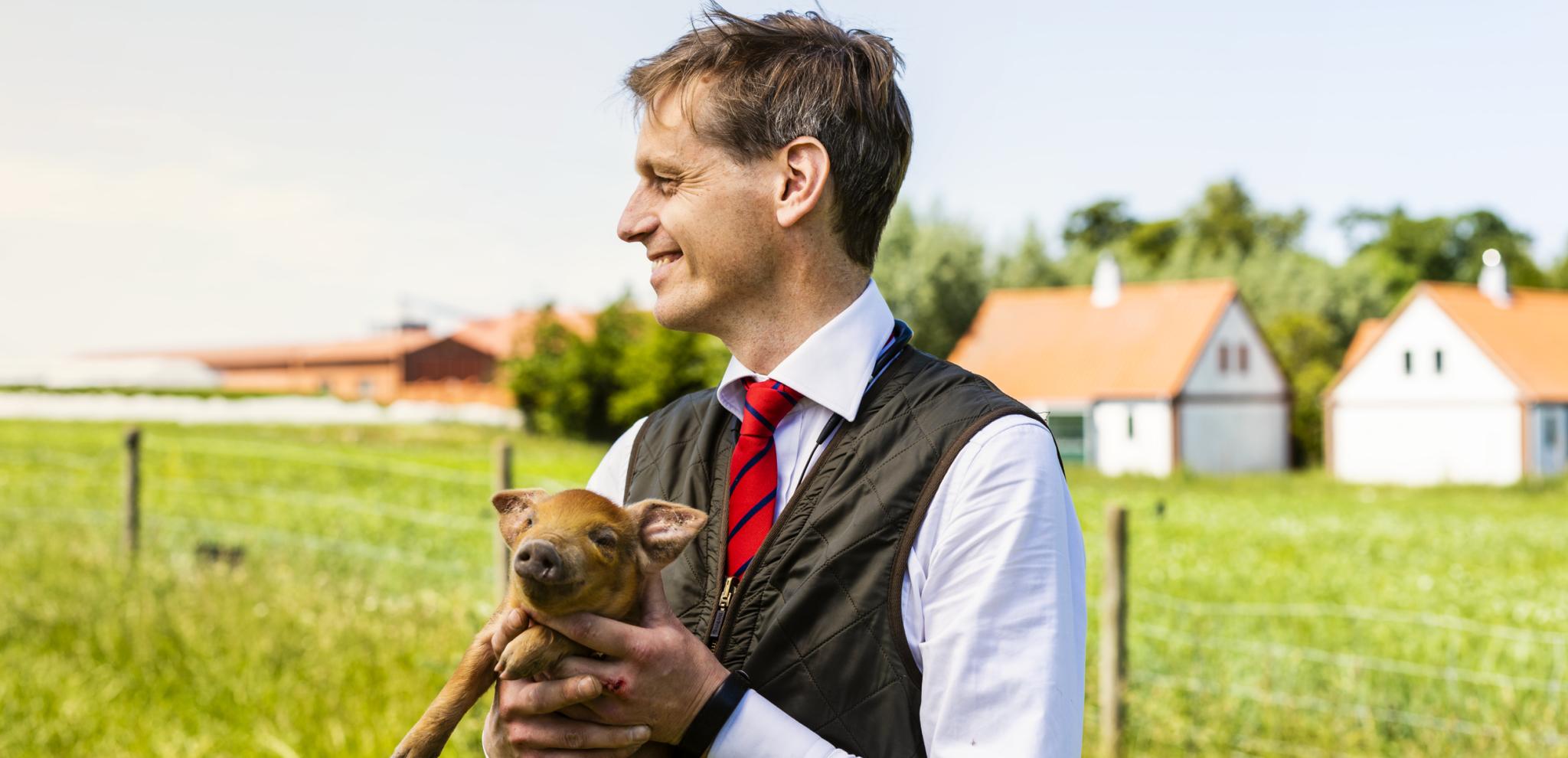 Smiling man in traditional vest and tie holding small pig outdoors with farm buildings and green fields in rural countryside background