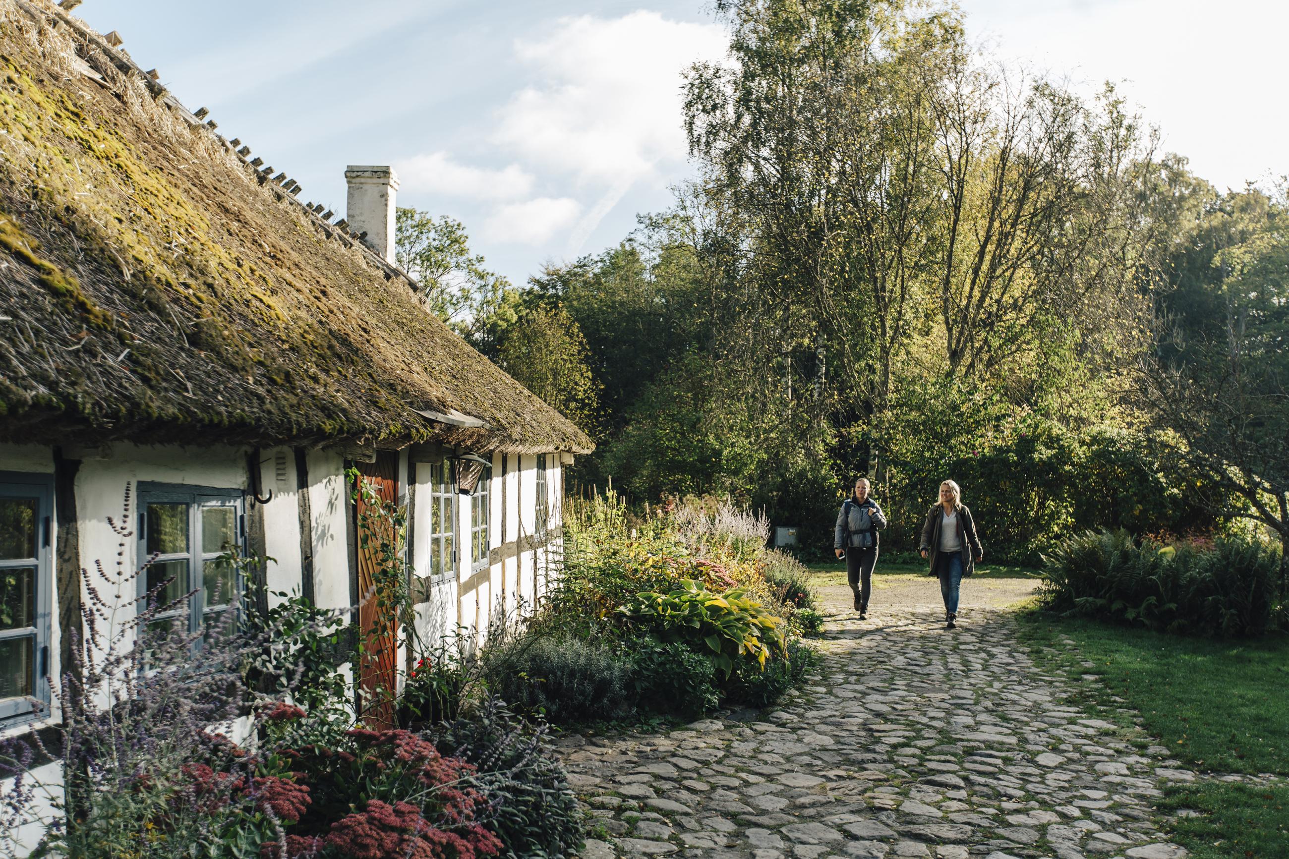 Zwei Frauen wandern auf einem Steinweg an einem denkmalgeschützten Fachwerkhaus vorbei.