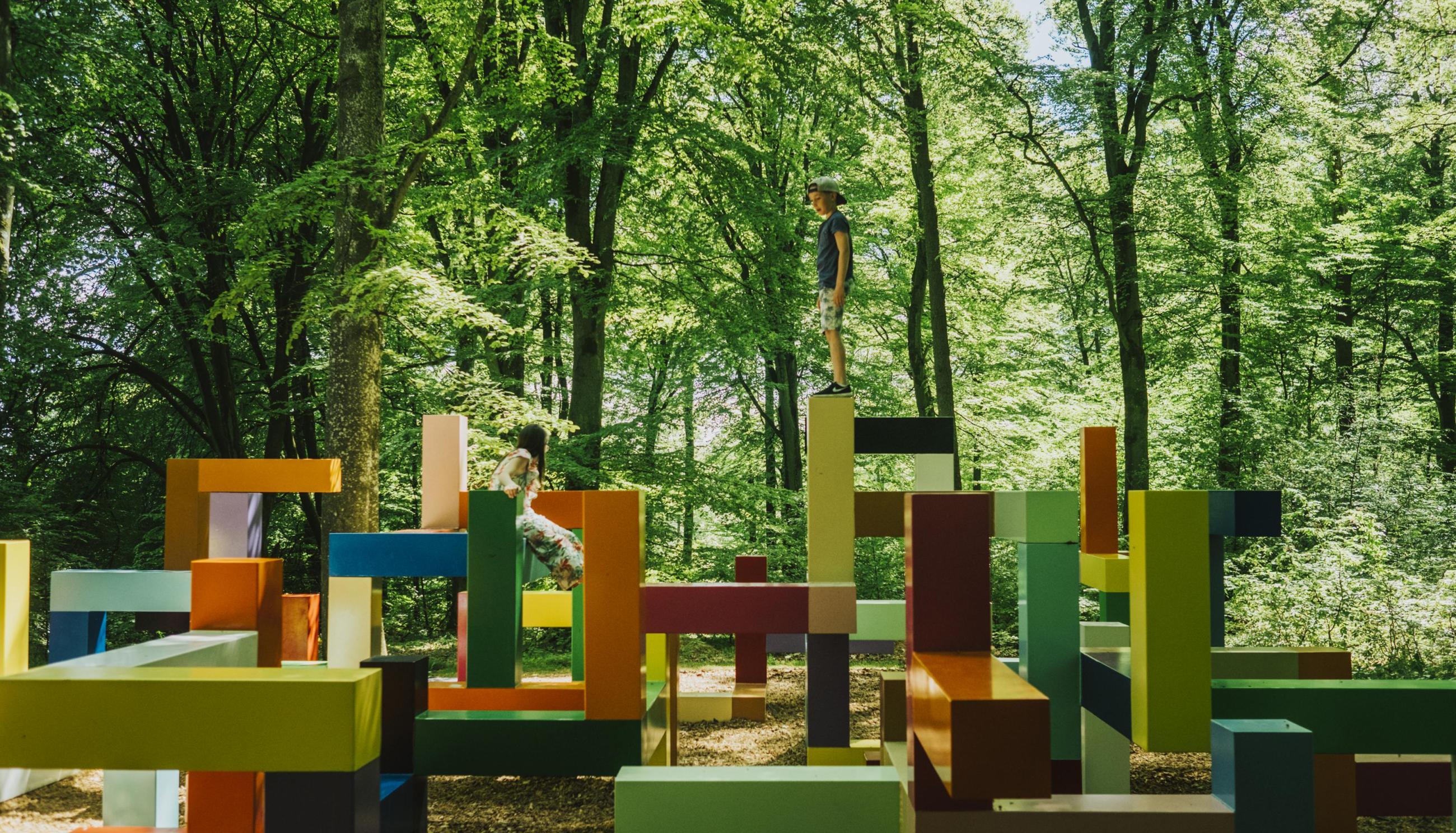 Kinder spielen auf einem mit abstrakten Formen gestalteten Spielplatz in Wanås, umgeben von hohen Bäumen.