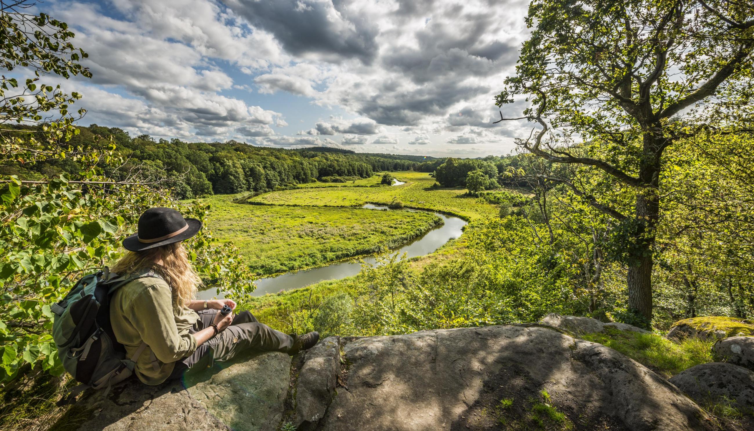 Eine Frau mit Hut sitzt auf einer Klippe und blickt auf ein grünes Flusstal.