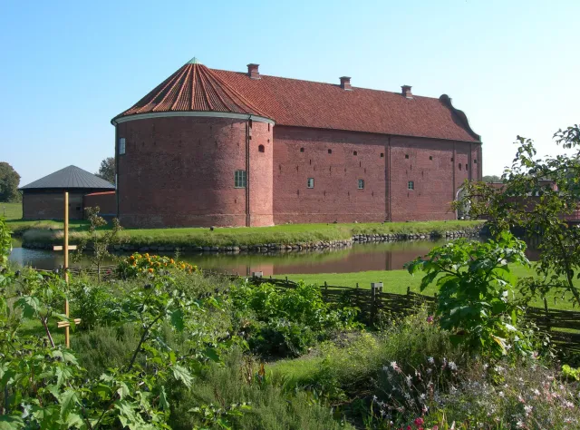 Tegelröd gammal stenbyggnad med ett runt torn och grön trädgårdsmiljö framför