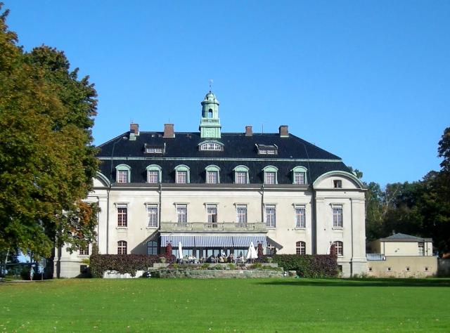 Weißes Schloss mit großer grüner Rasenfläche davor und blauem Himmel dahinter
