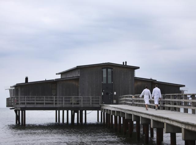 Open-air bathhouse with two people walking on a pier towards it 