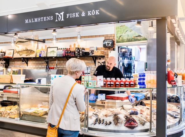 A woman shopping delicacies from a counter. A man helps her across the counter.