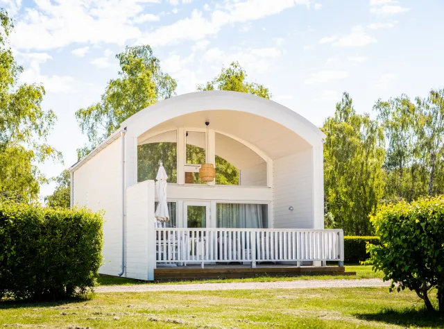 En vit stuga med välvt tak och terass med gräsmatta och buskar framför
