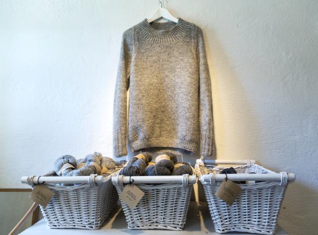 En ulltröja hänger på väggen med tre korgar under som innehåller ullgarn