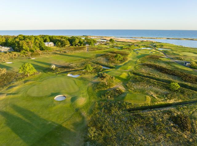 Luftaufnahme eines Küsten-Linksgolfplatzes mit welligen Fairways, Sandbunkern, Grüns und Meereshintergrund, Häuser zwischen Bäumen eingebettet