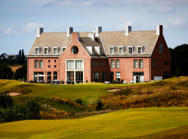 Stattliches Backsteinclubhaus auf gepflegtem Golfplatz mit markanten Dachgauben, Außensitzbereich, Sandbunkern und welligen Fairways im Vordergrund
