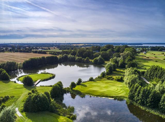Luftaufnahme eines Golfplatzes mit Wasserhindernissen, baumgesäumten Fairways und von See umgebenen Grüns, entfernten Windrädern und Landschaftspanorama