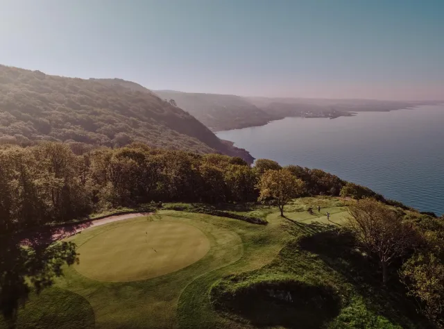 Erhöhte Ansicht eines dramatischen Klippen-Golflochs mit Grün über Küstenlinie, Golfspieler zwischen bewaldeten Hängen und weitem Meeresblick