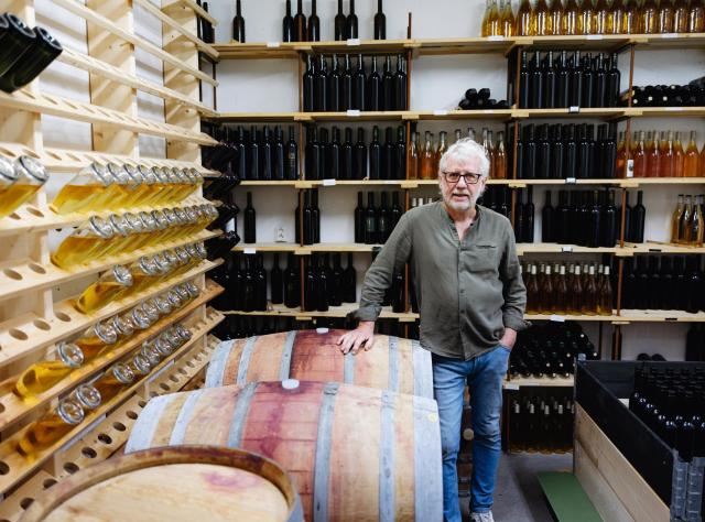 Winemaker with glasses standing beside wooden wine barrel in cellar, surrounded by shelves filled with wine bottles and wooden wine racks
