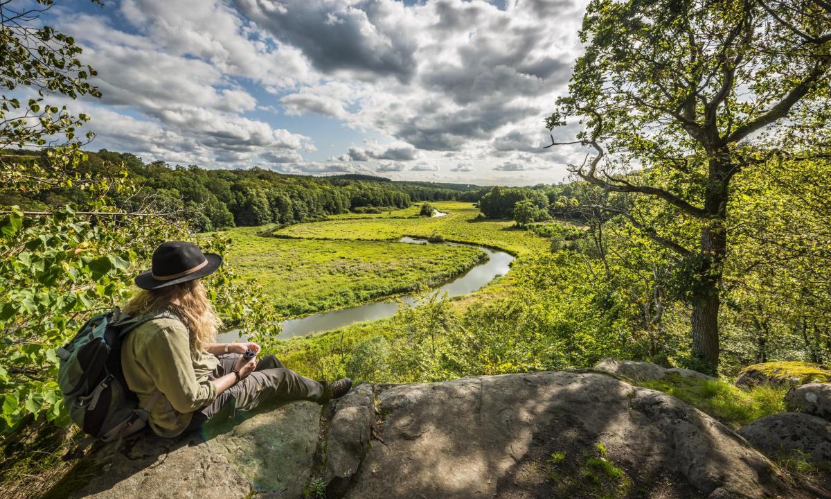 Eine Frau mit Hut sitzt auf einer Klippe und blickt auf ein grünes Flusstal.